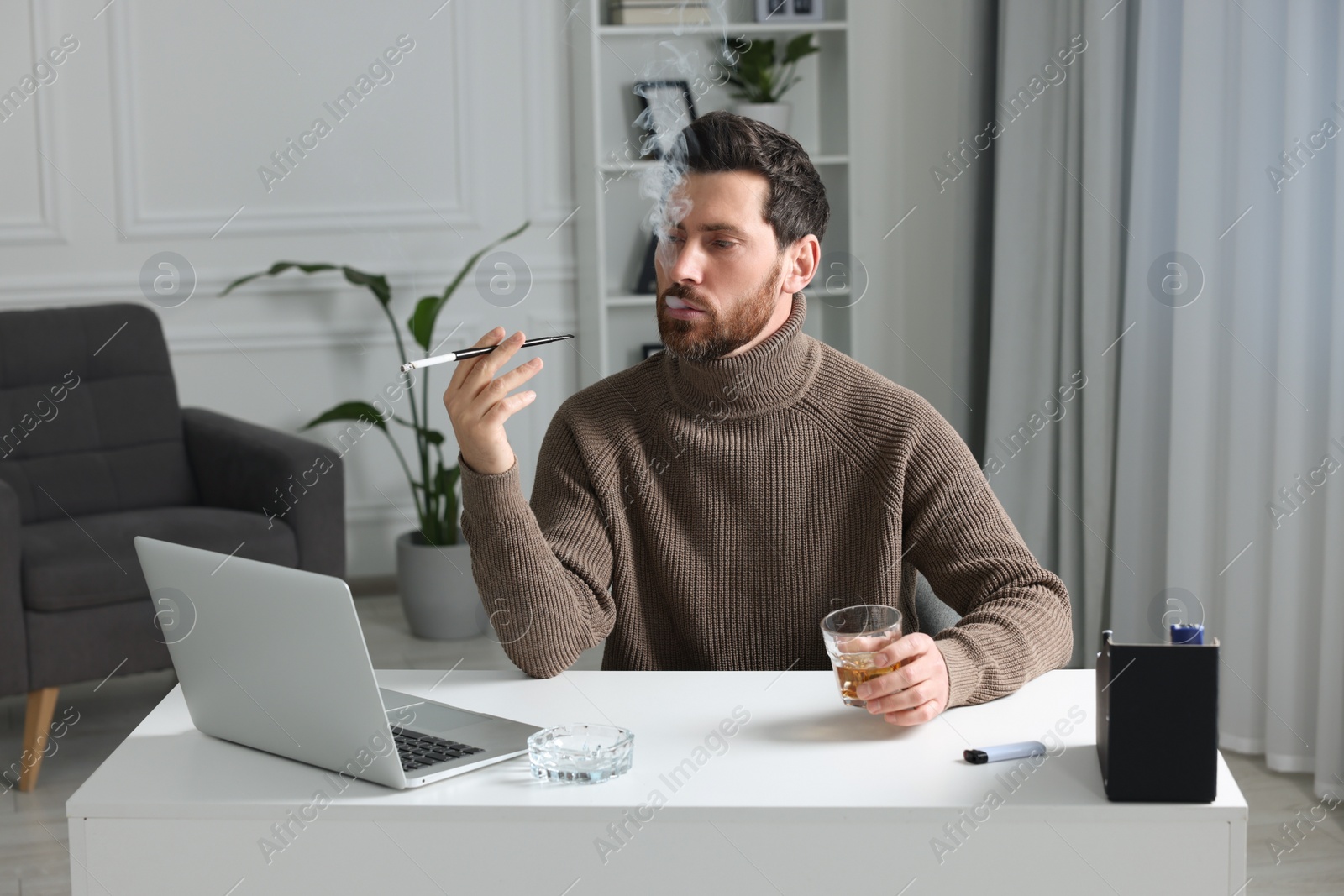 Photo of Man using long cigarette holder for smoking and holding glass of whiskey at workplace in office