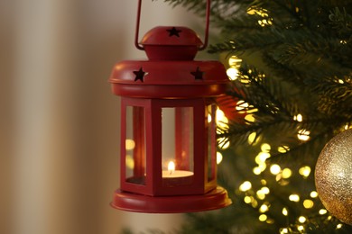 Photo of Christmas lantern with burning candle, festive lights and ball on fir tree against blurred background, closeup