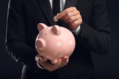 Photo of Businessman putting coin in piggy bank on black background, closeup