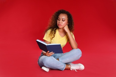 Beautiful African-American young woman reading book on red background