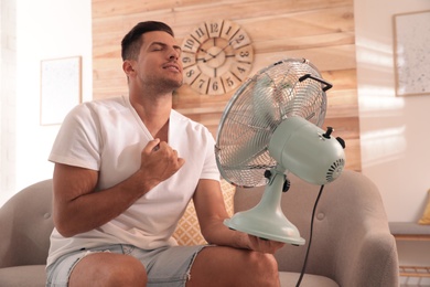 Photo of Man enjoying air flow from fan on sofa in living room. Summer heat