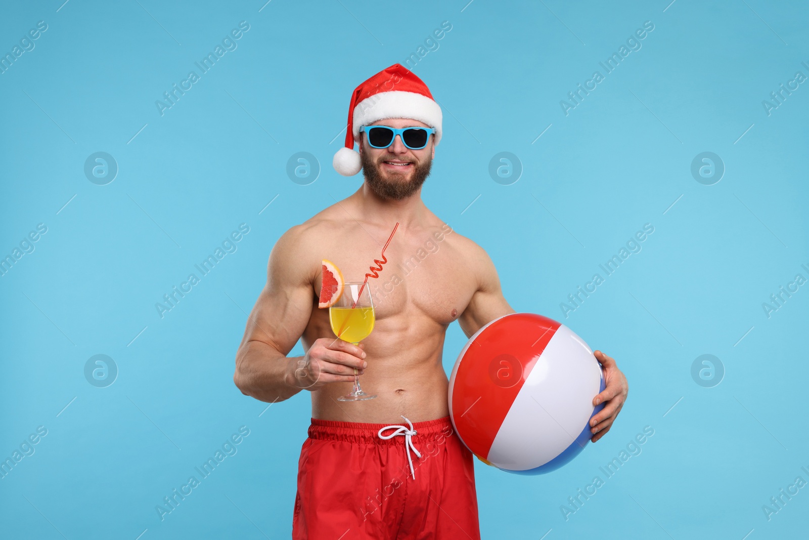 Photo of Attractive young man with muscular body in Santa hat holding ball and cocktail on light blue background