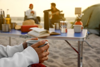 Friends resting near camping tent at beach, focus on woman with cup of hot drink