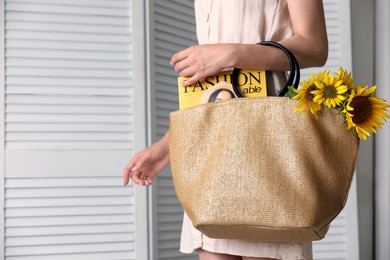 Photo of Woman holding beach bag with magazine and beautiful bouquet of sunflowers indoors, closeup