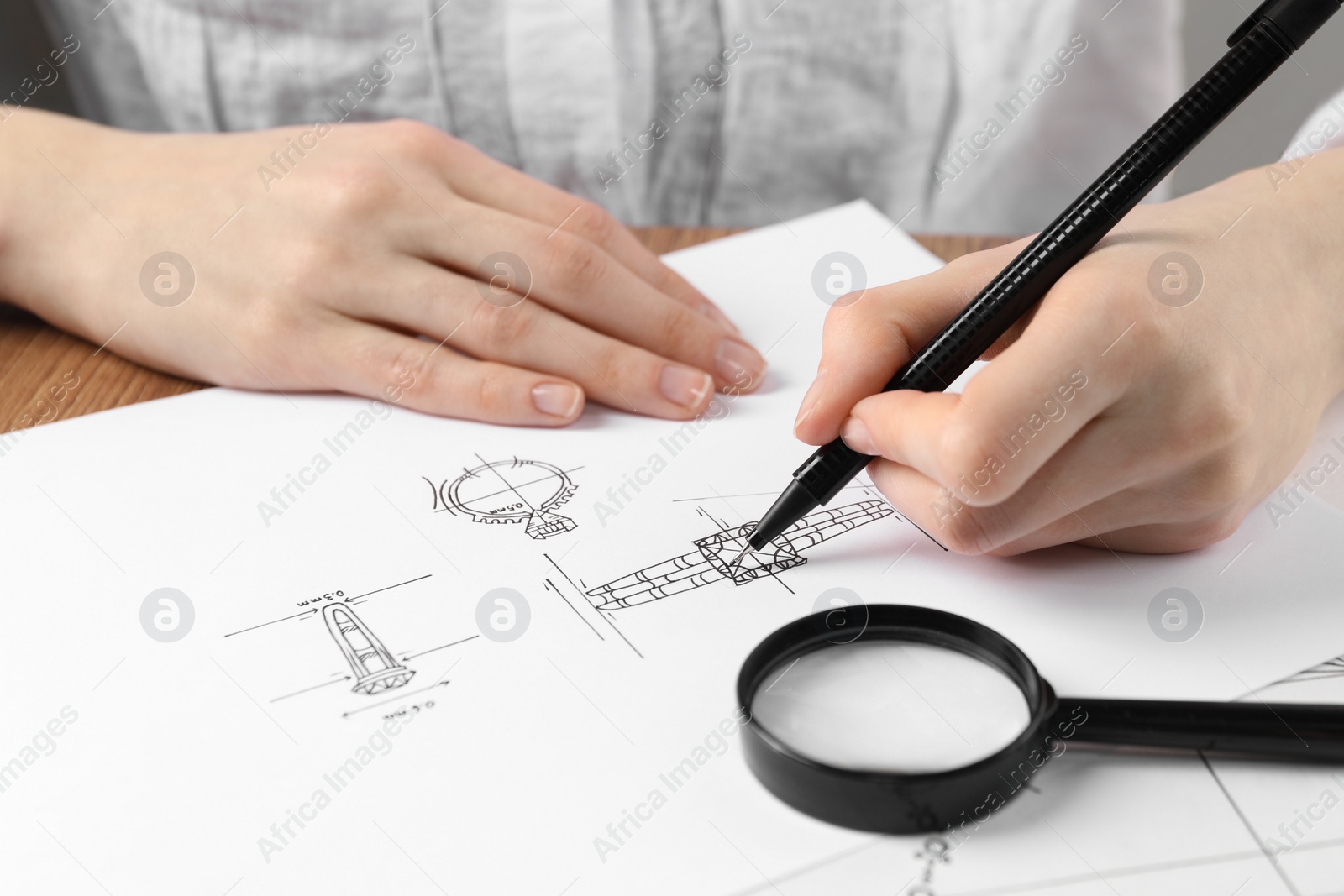 Photo of Jeweler drawing sketch of elegant ring on paper at wooden table, closeup