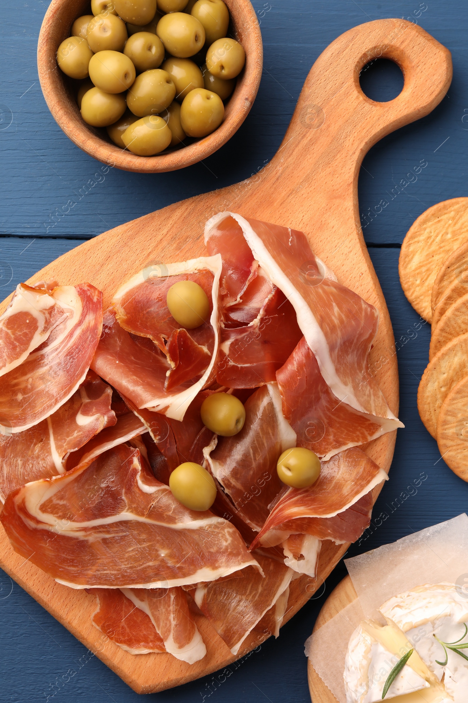 Photo of Slices of tasty cured ham, olives and cheese on blue wooden table, flat lay