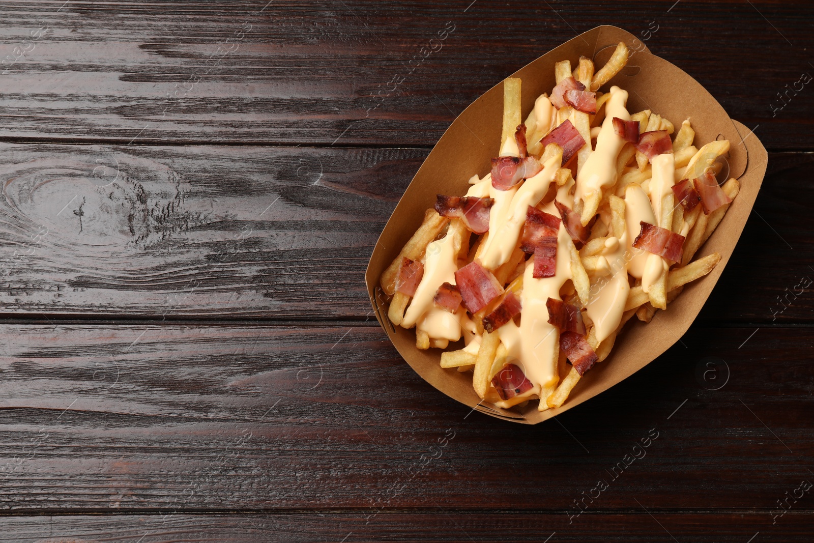 Photo of Tasty potato fries, cheese sauce and bacon in paper container on wooden table, top view. Space for text