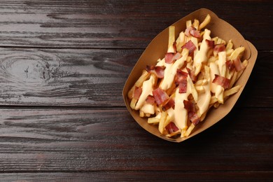 Tasty potato fries, cheese sauce and bacon in paper container on wooden table, top view. Space for text