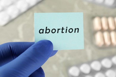 Photo of Doctor holding paper card with word ABORTION over table with pills, closeup
