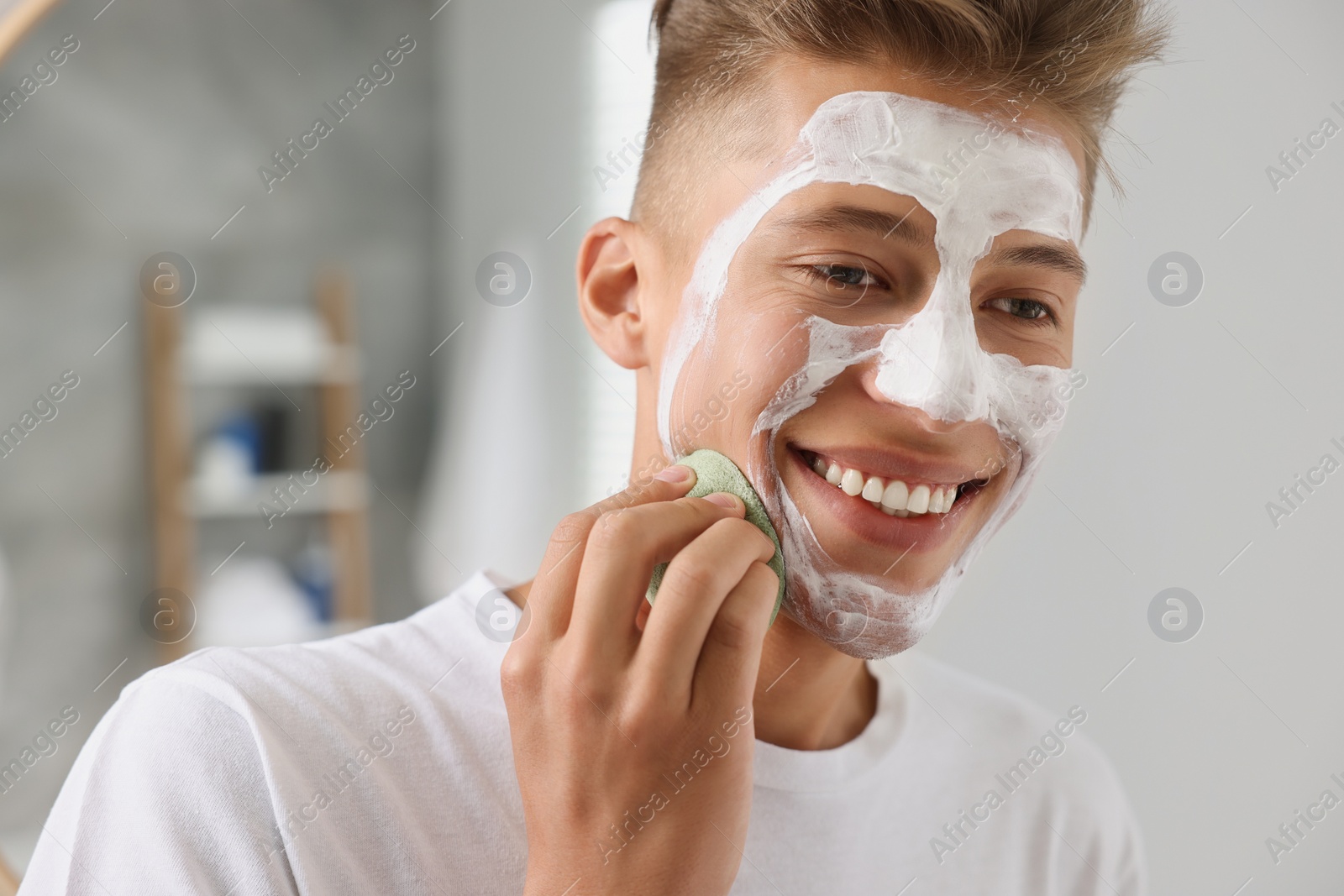Photo of Happy young man washing off face mask with sponge in bathroom