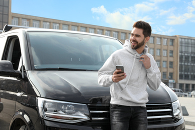 Handsome young man with smartphone near modern car outdoors
