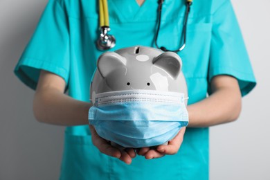 Photo of Doctor holding light gray ceramic piggy bank with mask against white wall, closeup. Medical insurance