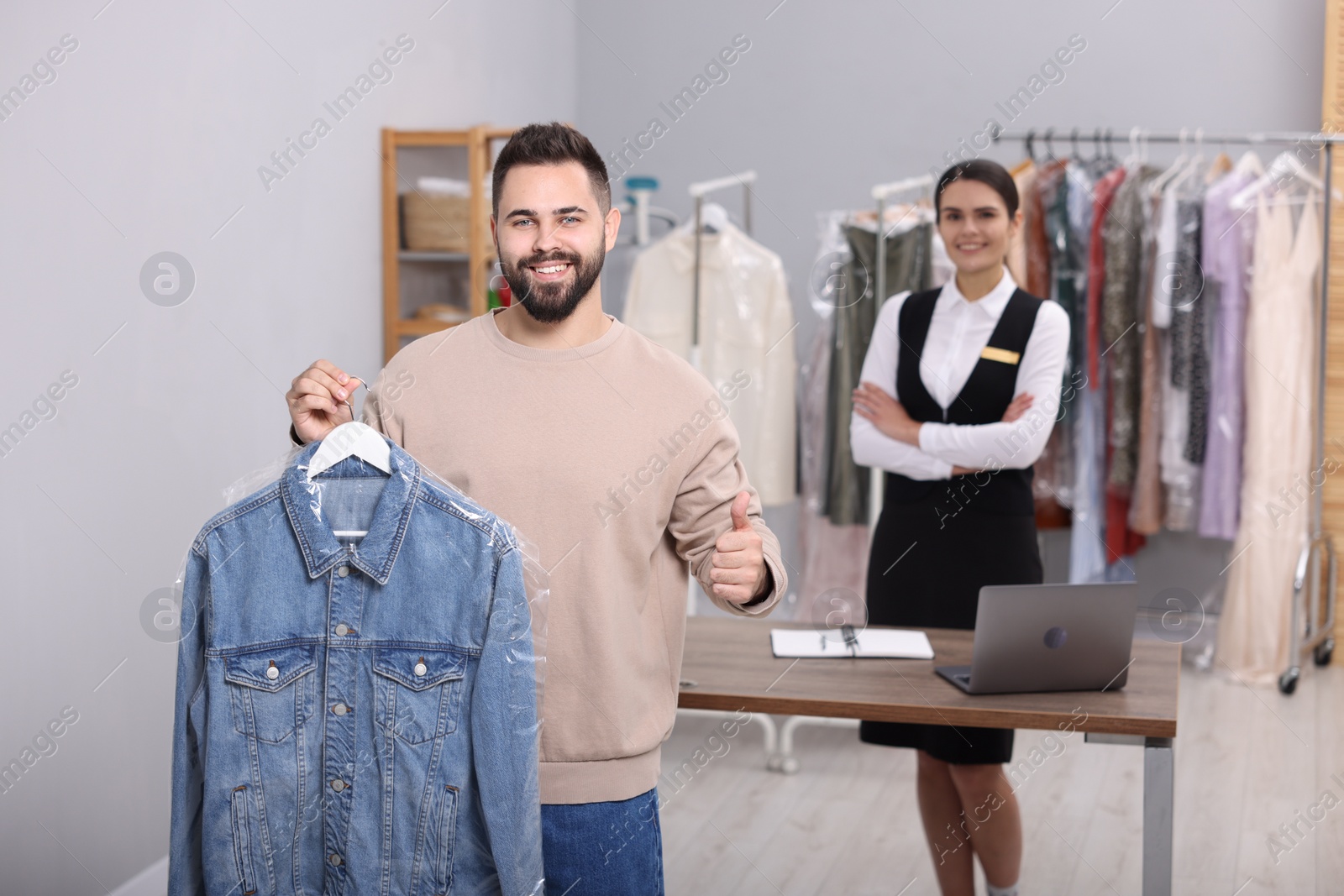 Photo of Dry-cleaning service. Man holding hanger with denim jacket and showing thumb up indoors. Happy worker at workplace