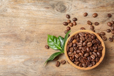 Bowl of coffee beans and fresh green leaves on wooden table, top view with space for text