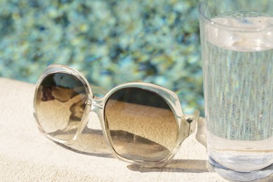 Stylish sunglasses and glass of water near outdoor swimming pool on sunny day, closeup
