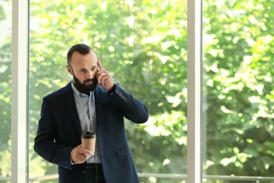 Photo of Portrait of mature man in elegant suit with coffee talking on mobile phone near window. space for text