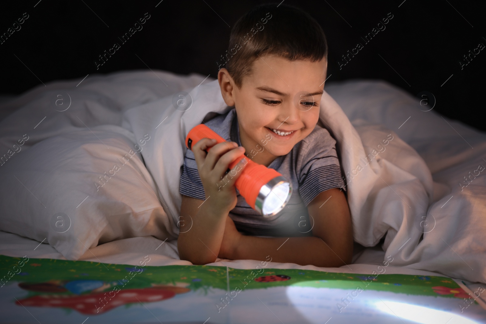 Photo of Little boy with flashlight reading book in bed at home