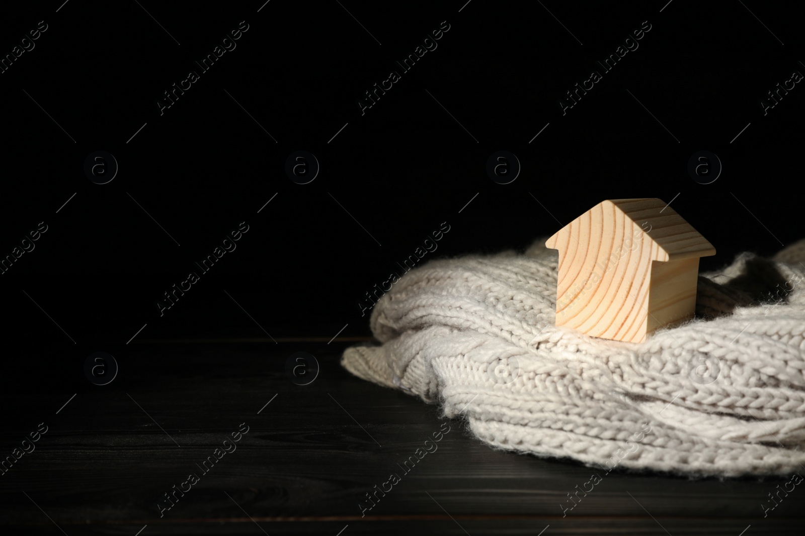 Photo of Wooden house model and scarf on table against black background, space for text. Heating efficiency