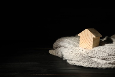 Photo of Wooden house model and scarf on table against black background, space for text. Heating efficiency