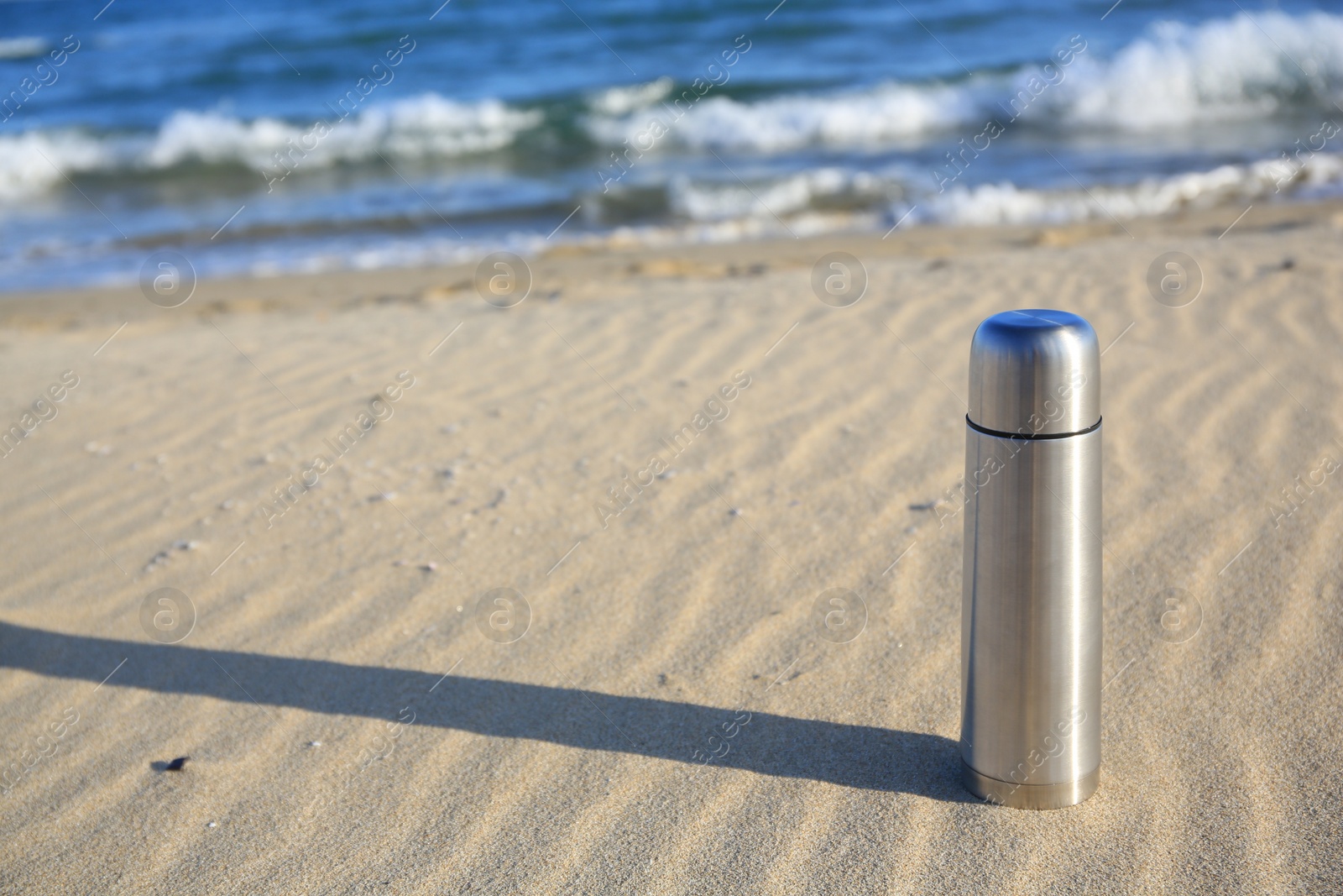 Photo of Metallic thermos with hot drink on sand near sea, space for text