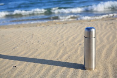 Metallic thermos with hot drink on sand near sea, space for text