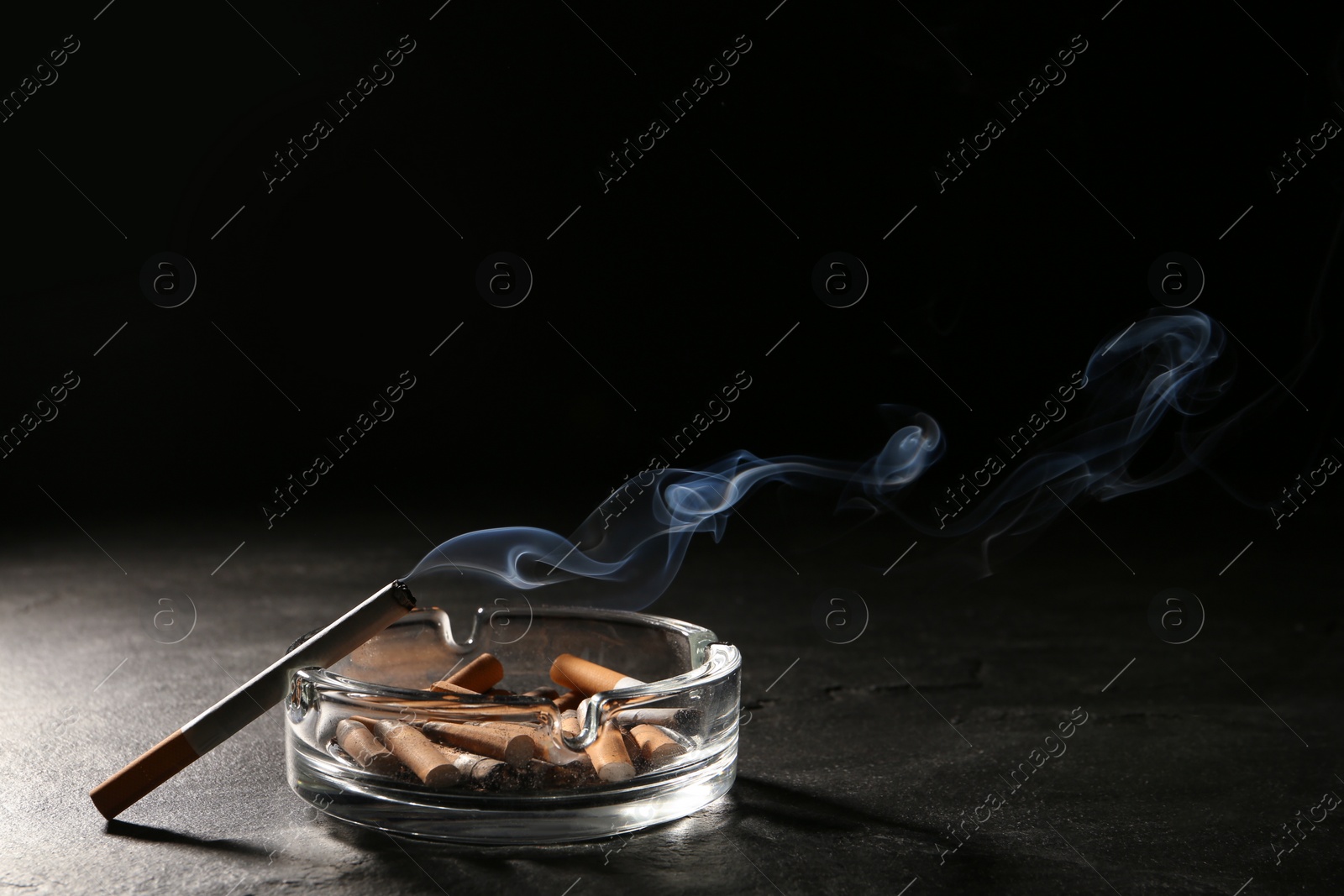 Photo of Glass ashtray with stubs and smoldering cigarette on grey table against black background. Space for text