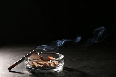 Glass ashtray with stubs and smoldering cigarette on grey table against black background. Space for text