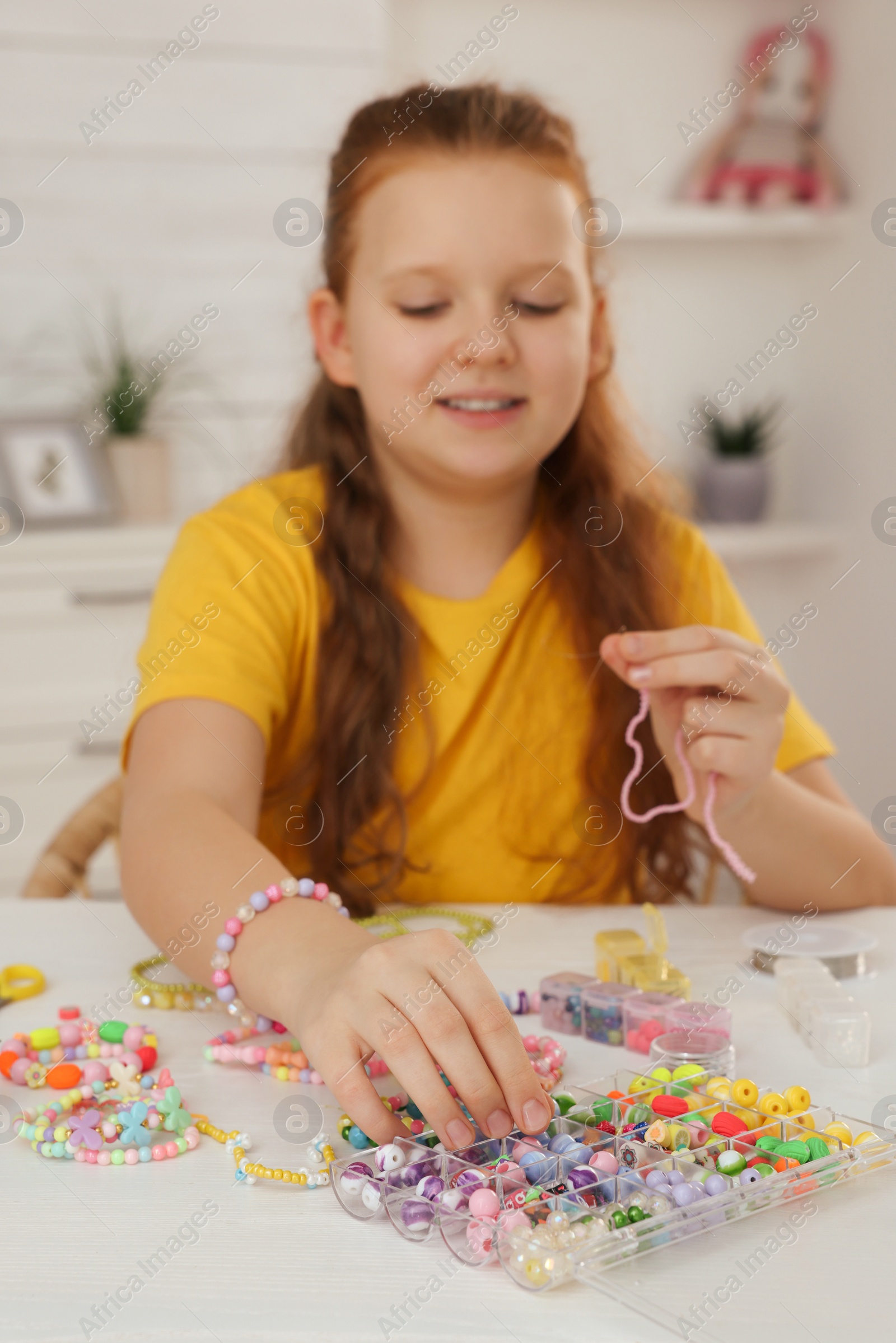 Photo of Cute girl making beaded jewelry at table in room, focus on hand