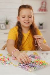 Photo of Cute girl making beaded jewelry at table in room, focus on hand