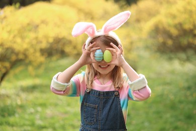 Photo of Easter celebration. Little girl in bunny ears covering eyes with painted eggs outdoors