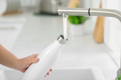 Photo of Woman pouring fresh water from tap into thermo bottle in kitchen, closeup