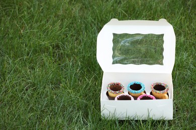 Photo of Box of delicious edible biscuit coffee cups decorated with sprinkles on green grass outdoors, space for text
