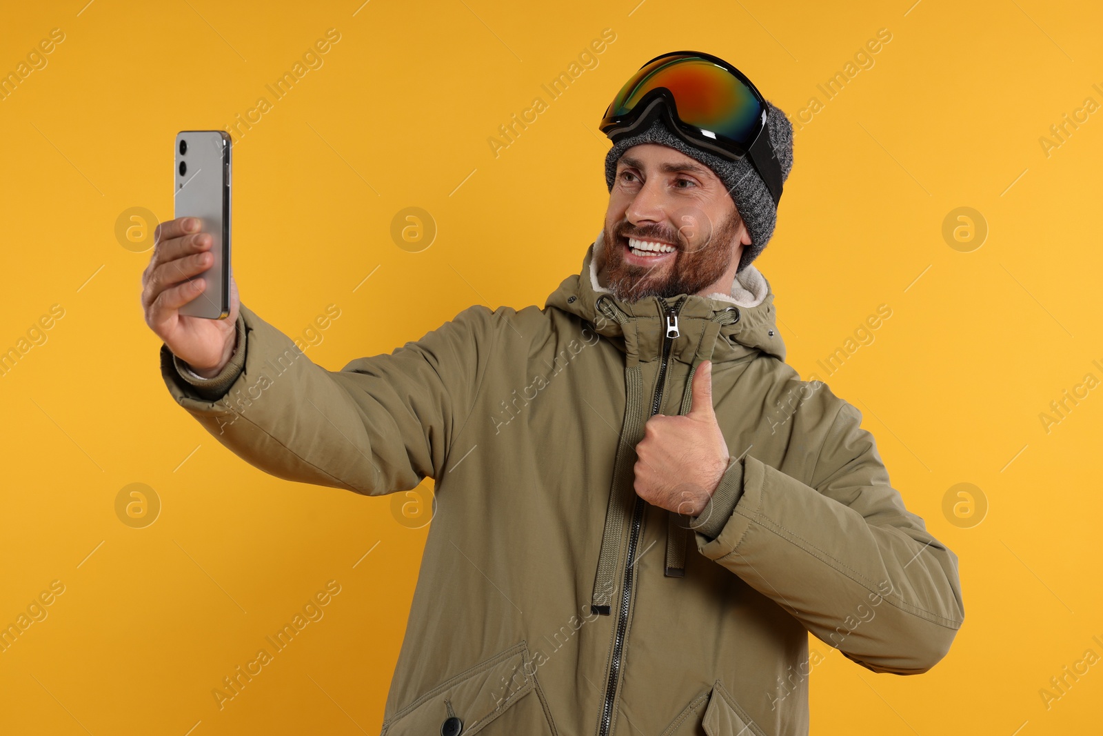 Photo of Winter sports. Happy man in ski suit and goggles taking selfie on orange background