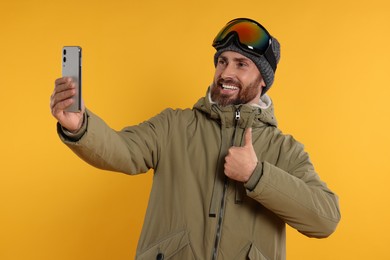 Photo of Winter sports. Happy man in ski suit and goggles taking selfie on orange background