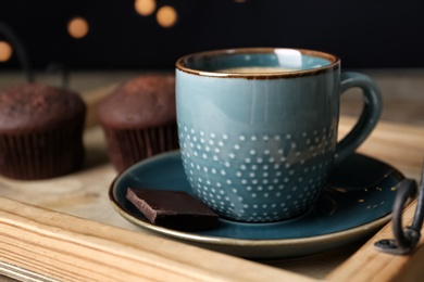 Photo of Cup with drink and piece of chocolate on wooden tray against blurred lights, closeup