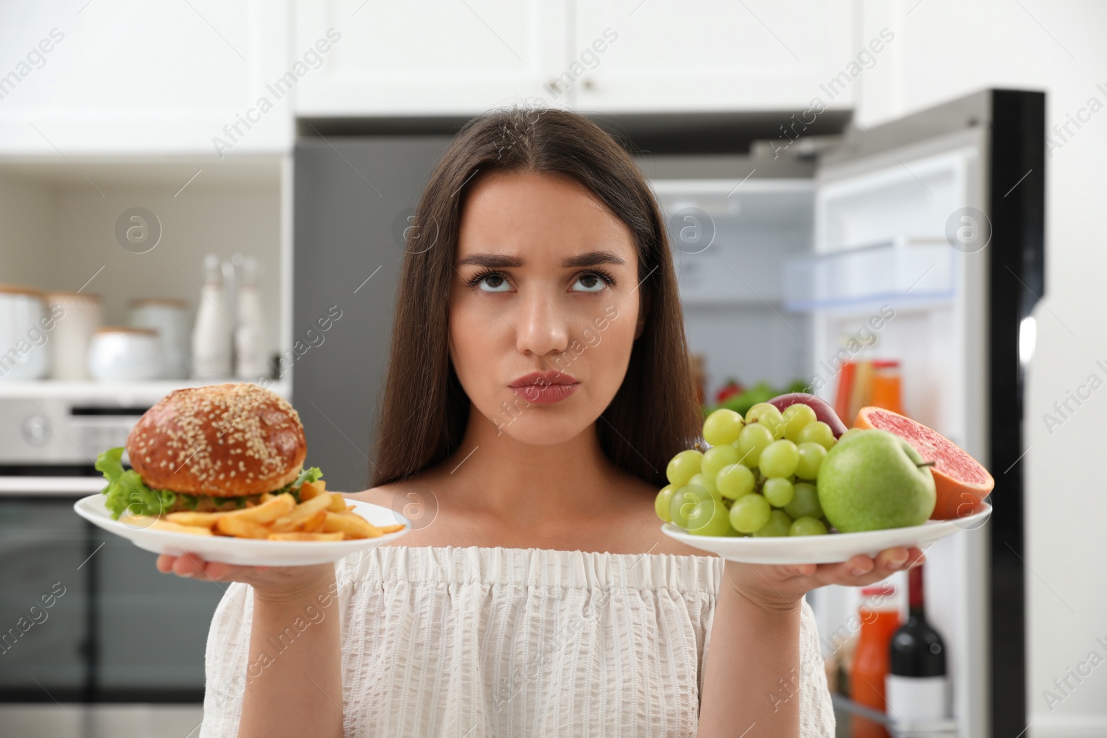 Photo of Woman choosing between fruits and burger with French fries near refrigerator in kitchen