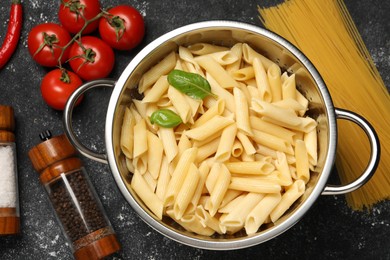 Photo of Cooked pasta in metal colander, products and spices on dark textured table, flat lay