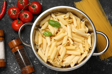 Cooked pasta in metal colander, products and spices on dark textured table, flat lay