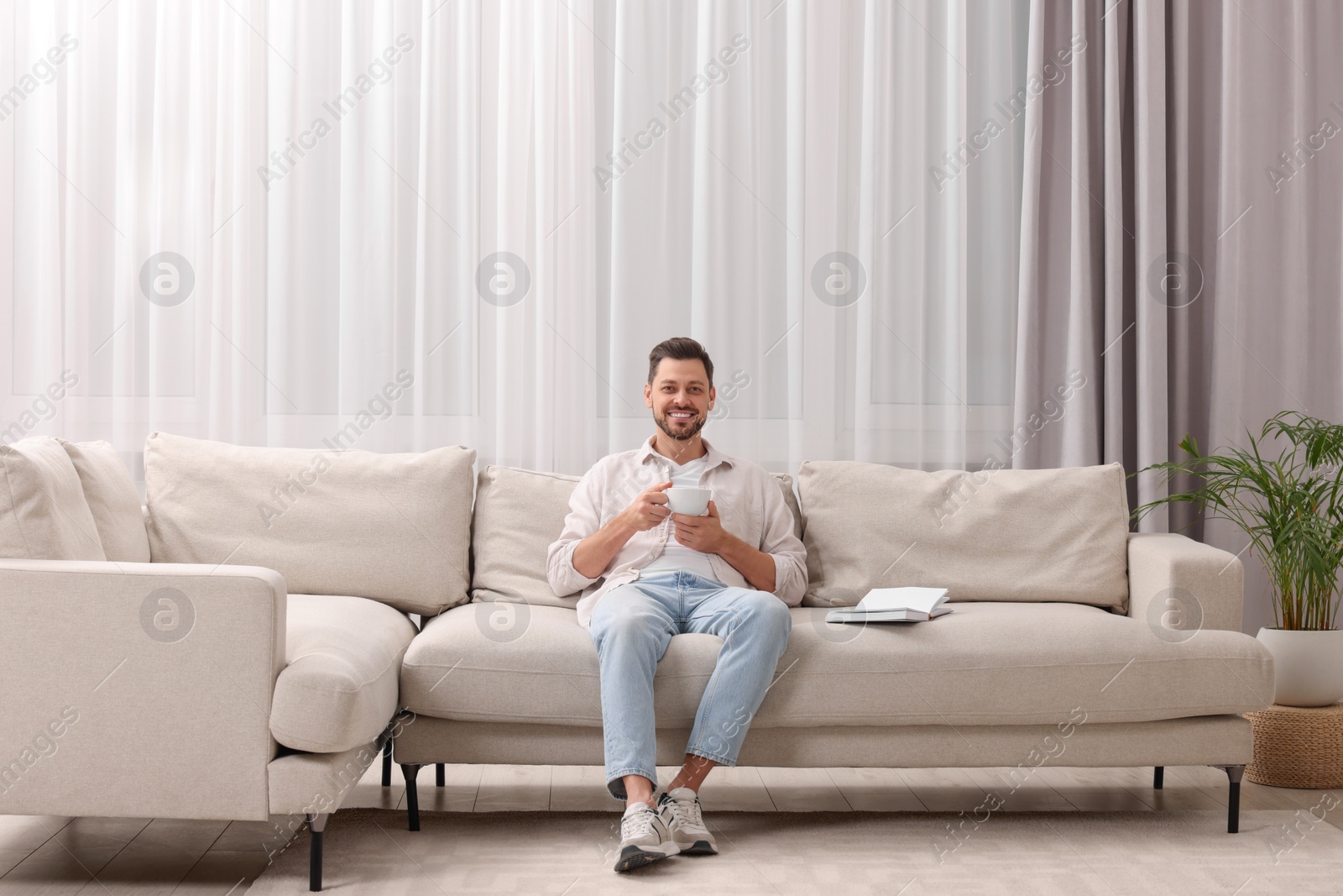 Photo of Happy man drinking coffee while resting on sofa near window with beautiful curtains in living room