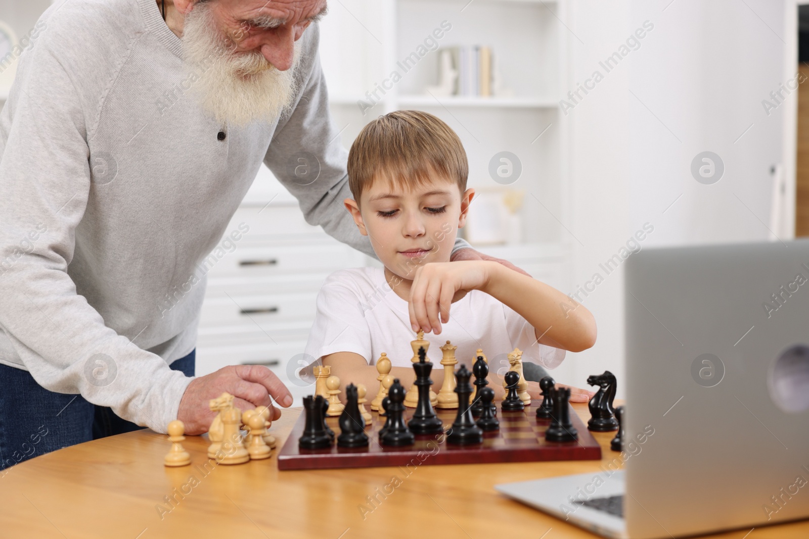 Photo of Grandfather teaching his grandson to play chess following online lesson at home