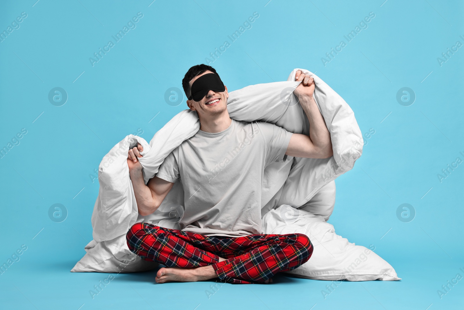 Photo of Happy man in pyjama and sleep mask wrapped in blanket on light blue background