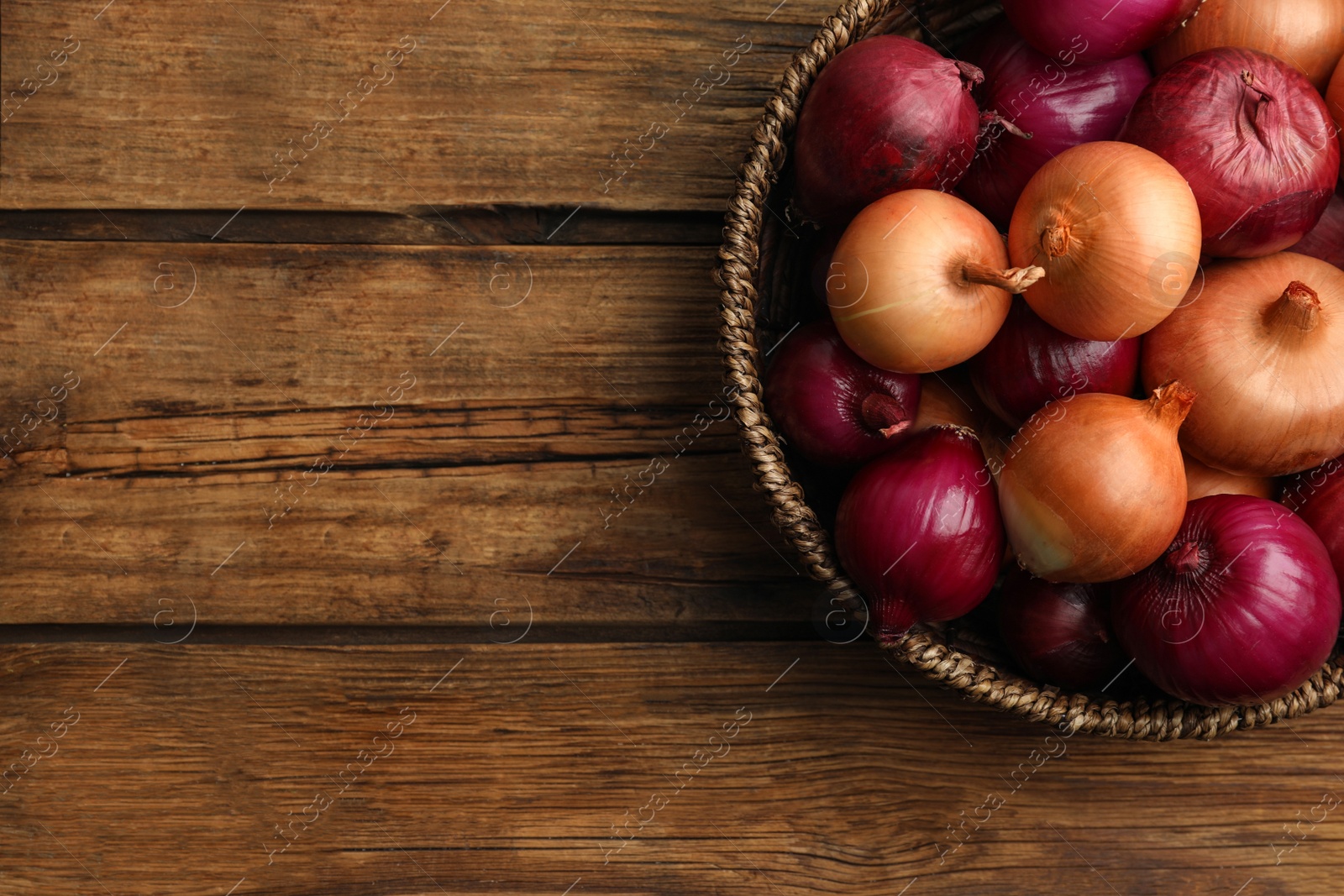 Photo of Onion bulbs in basket on wooden table, top view. Space for text