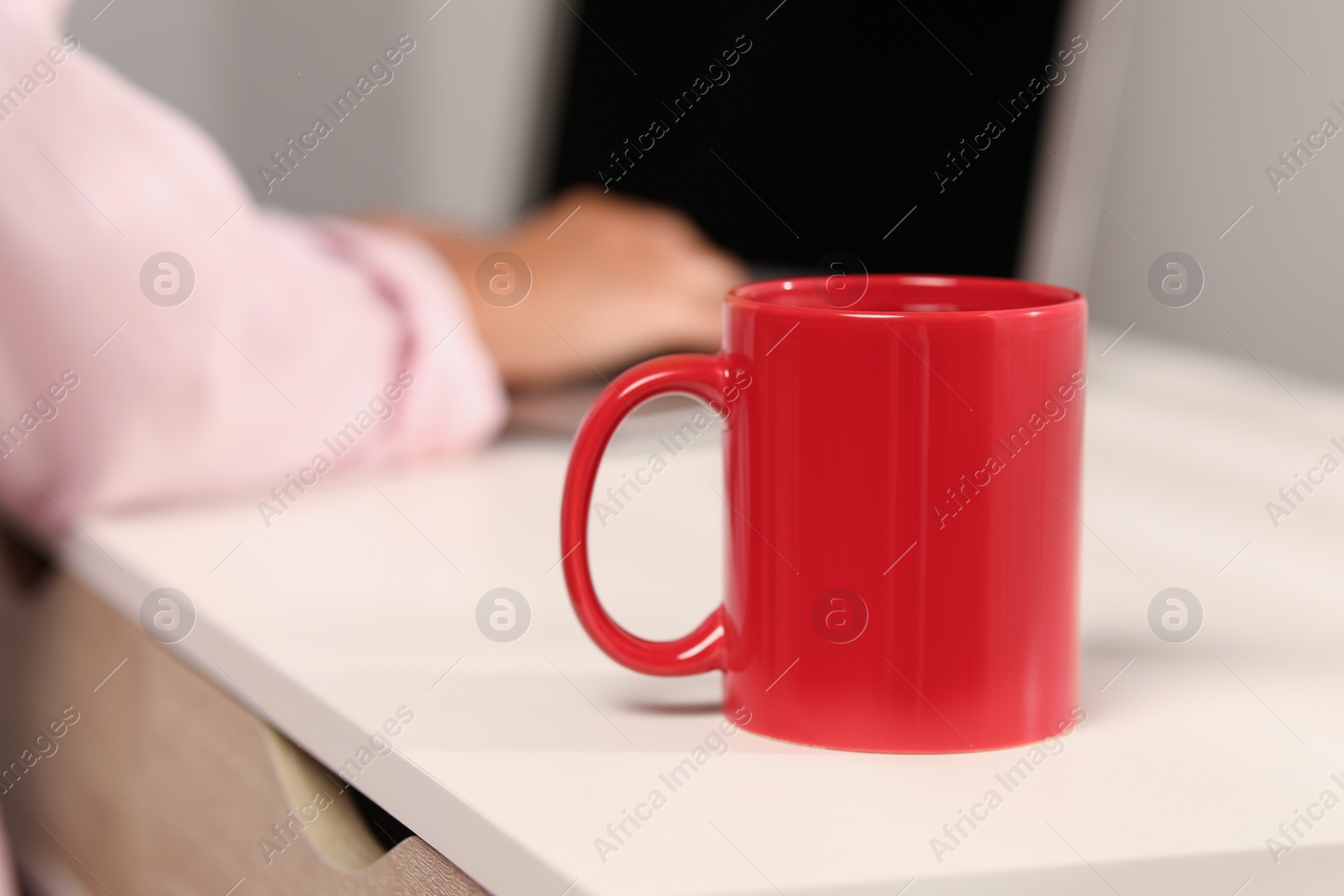 Photo of Red ceramic mug on white table, selective focus and space for text. Woman working with laptop at workplace, closeup