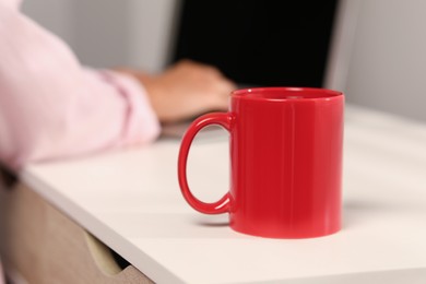 Photo of Red ceramic mug on white table, selective focus and space for text. Woman working with laptop at workplace, closeup