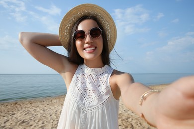 Beautiful young woman with straw hat and sunglasses taking selfie on beach. Stylish headdress