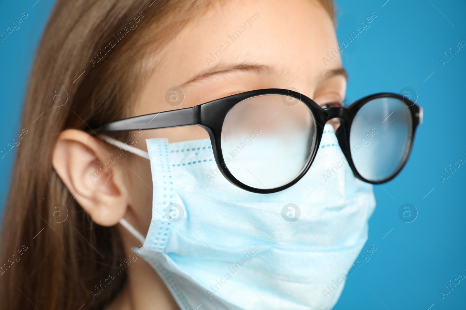 Photo of Little girl with foggy glasses caused by wearing disposable mask on blue background, closeup. Protective measure during coronavirus pandemic