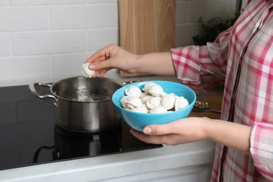 Woman putting frozen dumplings into saucepan with boiling water on cooktop in kitchen, closeup