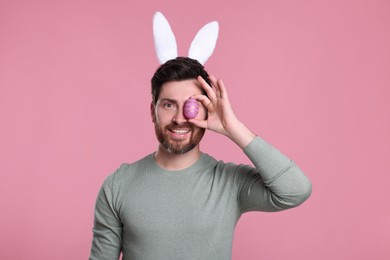 Happy man in cute bunny ears headband covering eye with Easter egg on pink background