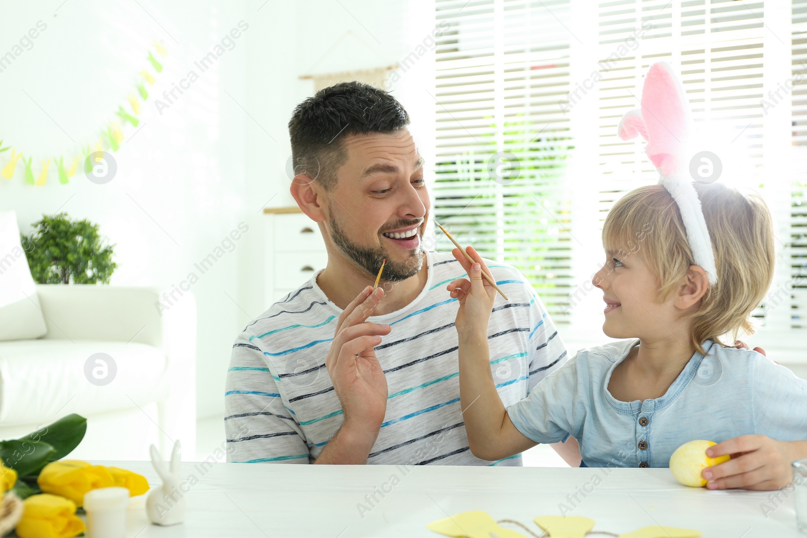 Photo of Happy son with bunny ears headband and his father having fun while painting Easter egg at home