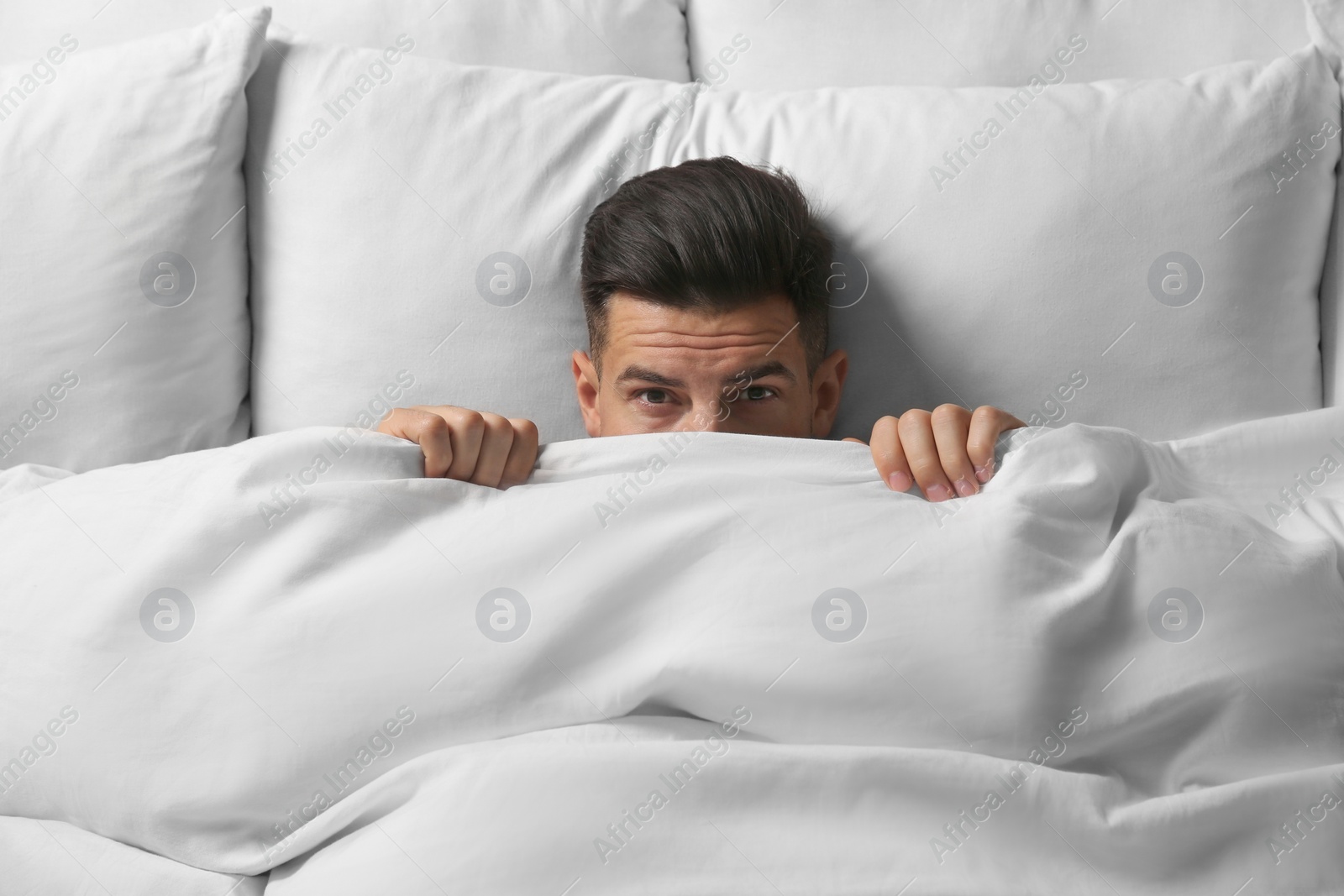 Photo of Handsome man covering his face with blanket while lying in bed at home